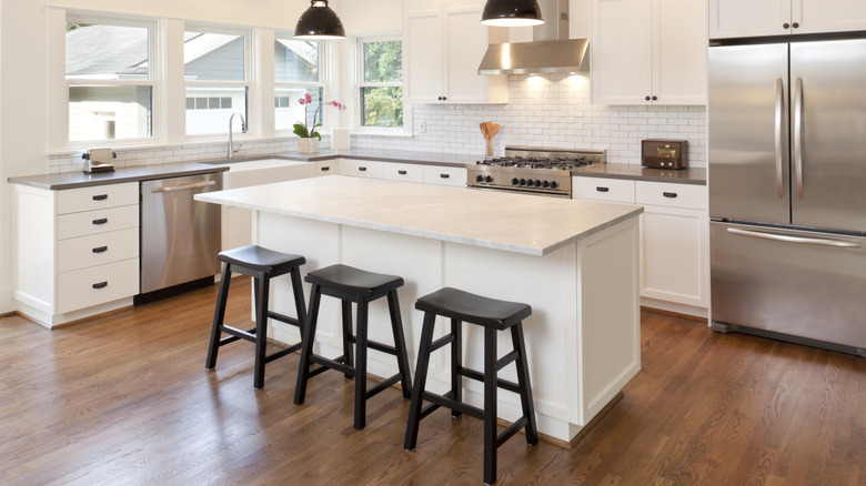 Nhà bếp hình chữ L với sàn gỗ, tủ màu trắng, thiết bị bằng thép không gỉ và bàn đảo với ba chiếc ghế đẩu màu đen