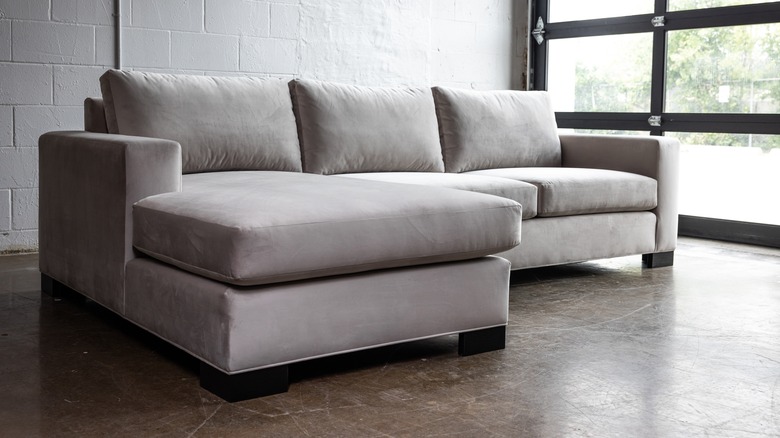 Ghế sofa nhung màu xám với chỗ ngồi lớn trong một không gian sống công nghiệp với các bức tường Cinderblock trắng, sàn bê tông và một cửa kính trên cao.