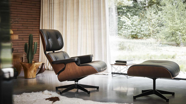 Phòng khách với một bản sao của Lounge da màu đen và gỗ Eames và Ottoman trước một bức tường gạch lộ ra và cửa sổ từ sàn đến trần với màn treo màu trắng
