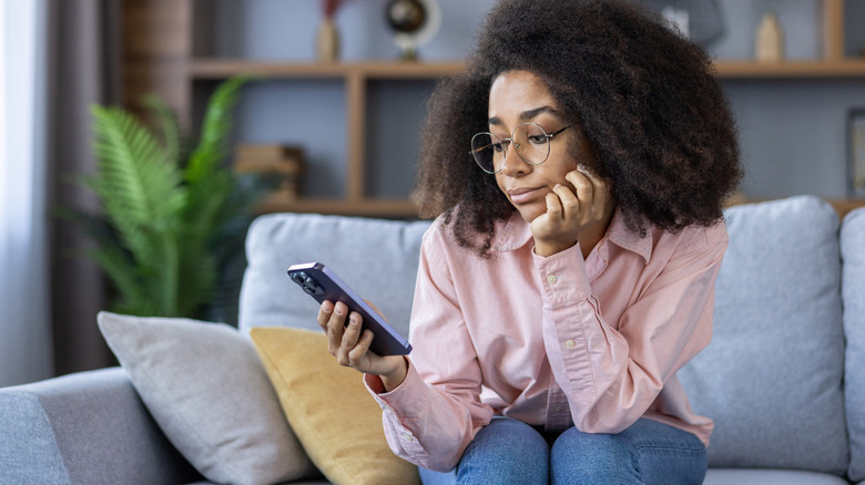 Người phụ nữ ngồi trên phòng khách sofa nhìn vào điện thoại của cô ấy với vẻ mặt chán nản trên khuôn mặt
