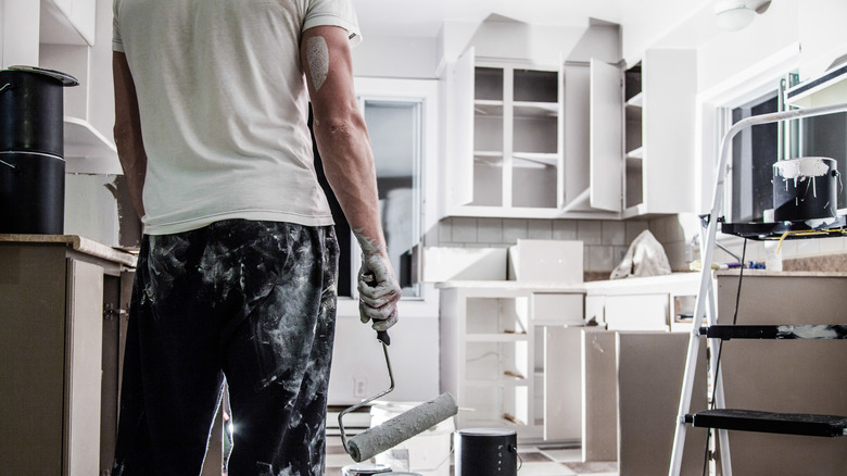 mặt sau của một người đàn ông cầm một con lăn sơn và phủ lên sơn với một nhà bếp còn dang dở trong nền