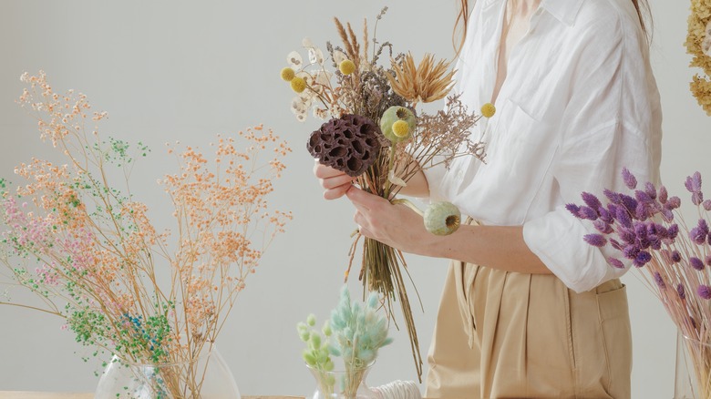 Một người phụ nữ với một bó hoa khô trong tay và nhiều bó hoa khô khác nhau trên bàn.
