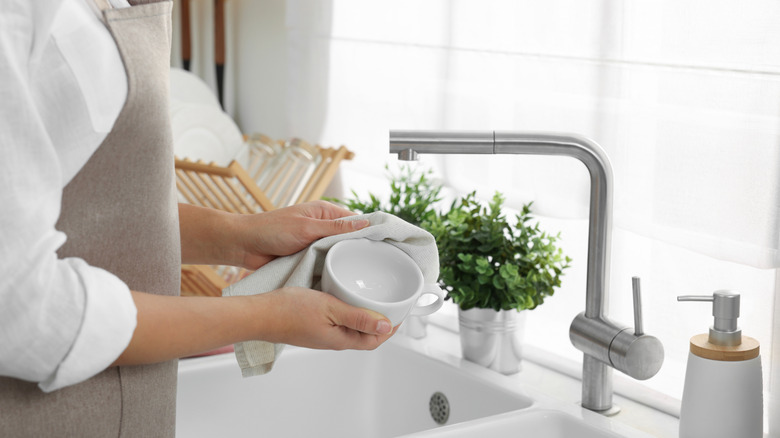 Một người phụ nữ đang lau cốc cà phê trắng bằng khăn lau bát đĩa màu trắng trên bồn rửa bát