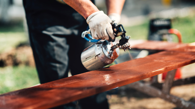 Người đàn ông đeo găng tay phun sơn lên gỗ chưa hoàn thiện ở ngoài trời cho dự án DIY