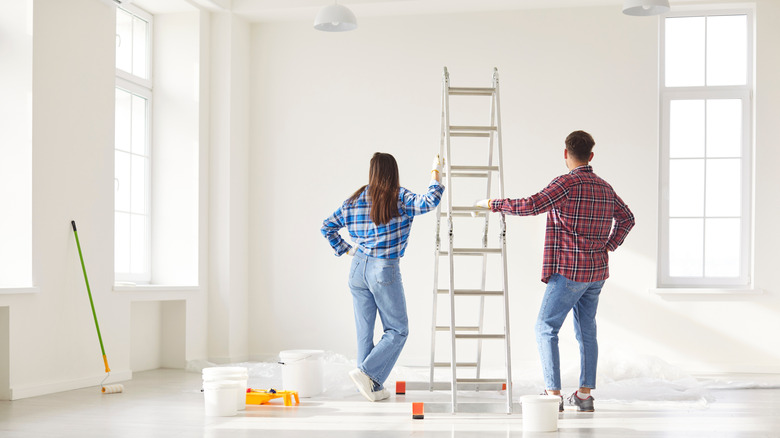 Một cặp vợ chồng trẻ đang nhìn vào bức tường trắng với những dụng cụ vẽ xung quanh họ